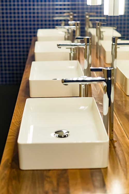 Commercial bathroom sinks with modern faucets arranged in a row on a wooden countertop, emphasizing plumbing installation services for businesses.