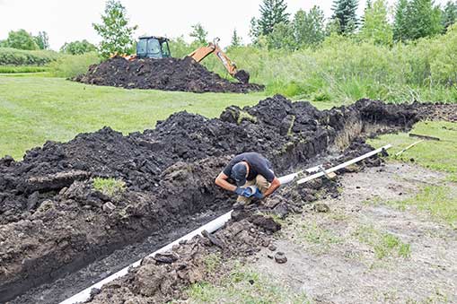 Excavation site with worker inspecting plumbing pipe in trench, heavy machinery and mounded soil in background, emphasizing proper excavation for plumbing systems.