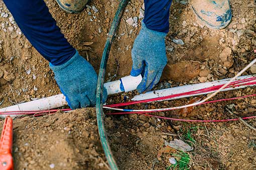 Hands wearing blue gloves working on plumbing pipes in an excavation site, surrounded by dirt and utility lines, illustrating preparation for plumbing excavation services by Leto Plumbing & Heating Inc.